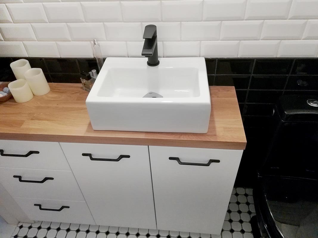 Biała, kwadratowa umywalka nablatowa z czarną armaturą, osadzona na drewnianym blacie i białej szafce z czarnymi uchwytami, w łazience z czarno-białymi płytkami.