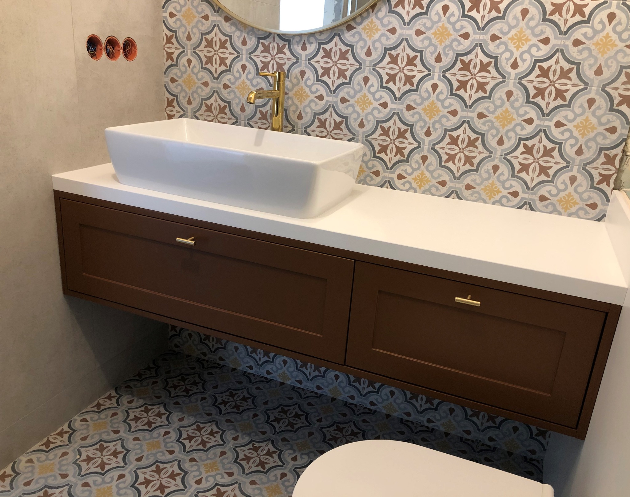 Biała, prostokątna umywalka nablatowa z złotą baterią, osadzona na brązowej szafce z dwoma szufladami i białym blatem, na tle wzorzystych płytek ściennych i podłogowych.