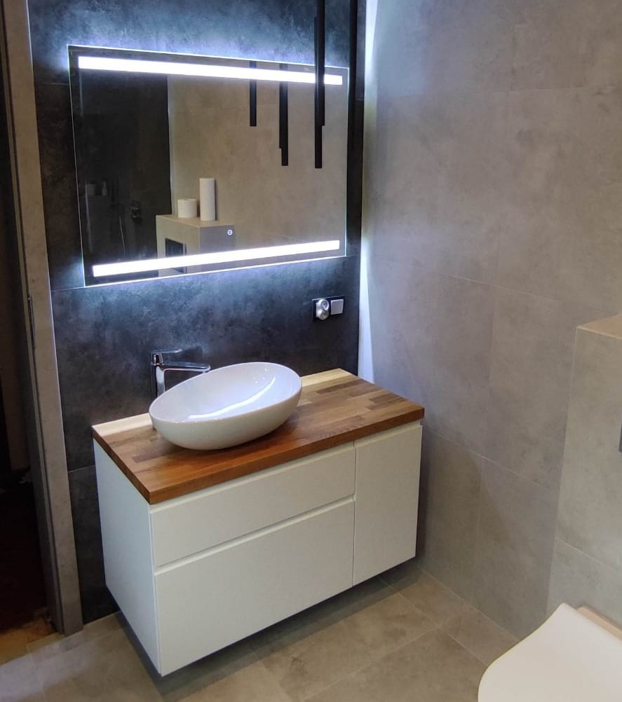 Nowoczesna łazienka z białą szafką pod umywalkę z drewnianym blatem, owalną umywalką nablatową i podświetlanym lustrem z czarnymi elementami dekoracyjnymi.