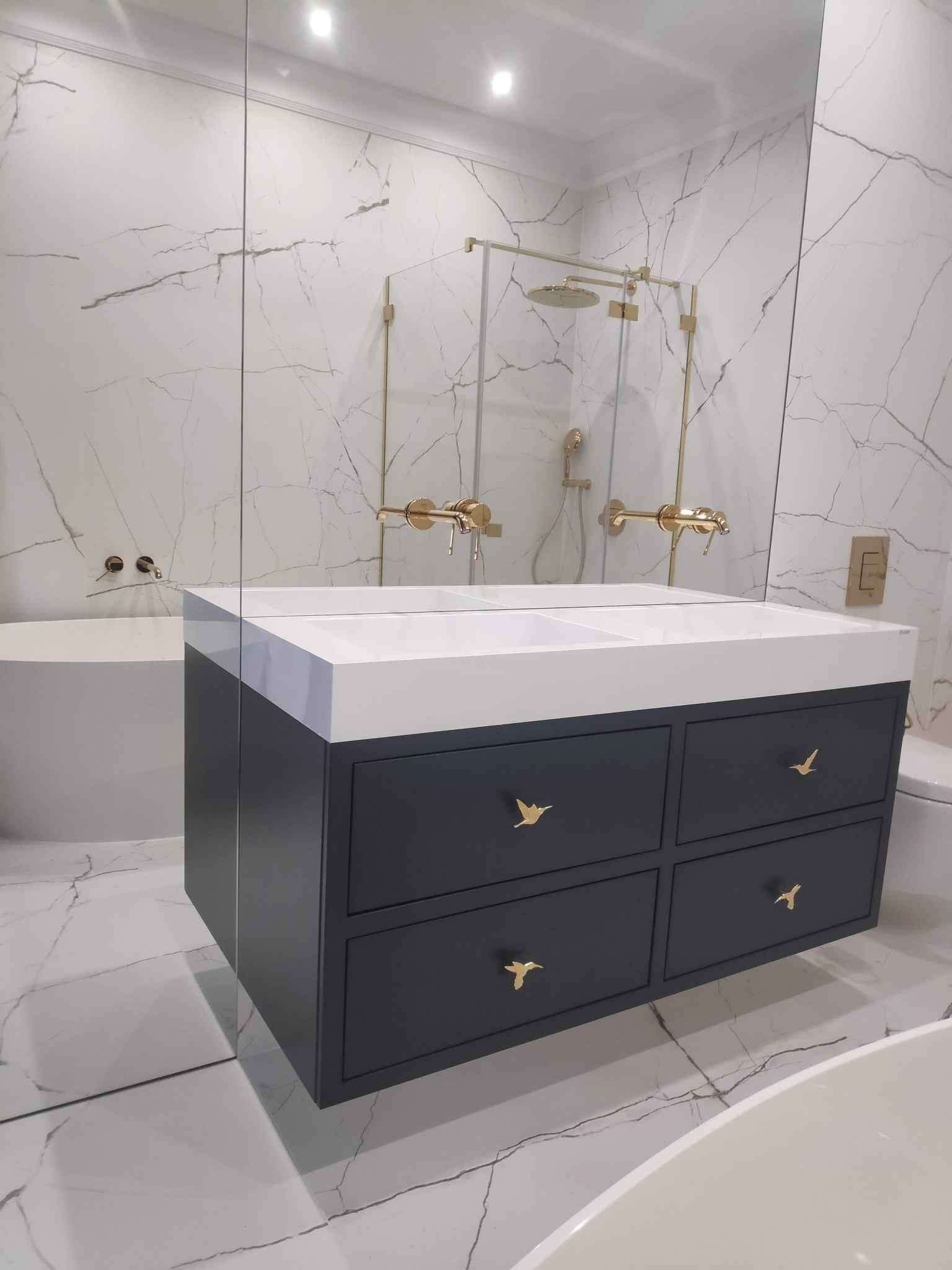 Nowoczesna łazienka z szarą szafką pod umywalkę z białym blatem i złotymi uchwytami w kształcie ptaków, marmurowe ściany i podłoga.