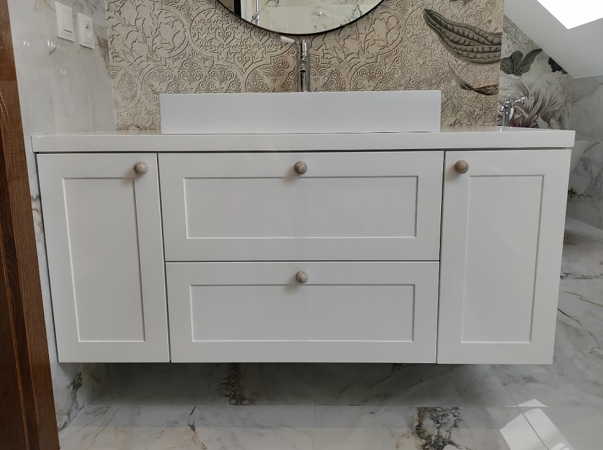Biała szafka łazienkowa pod umywalką z szufladami i drzwiczkami, okrągłe uchwyty, marmurowa podłoga i wzorzysta tapeta w tle.