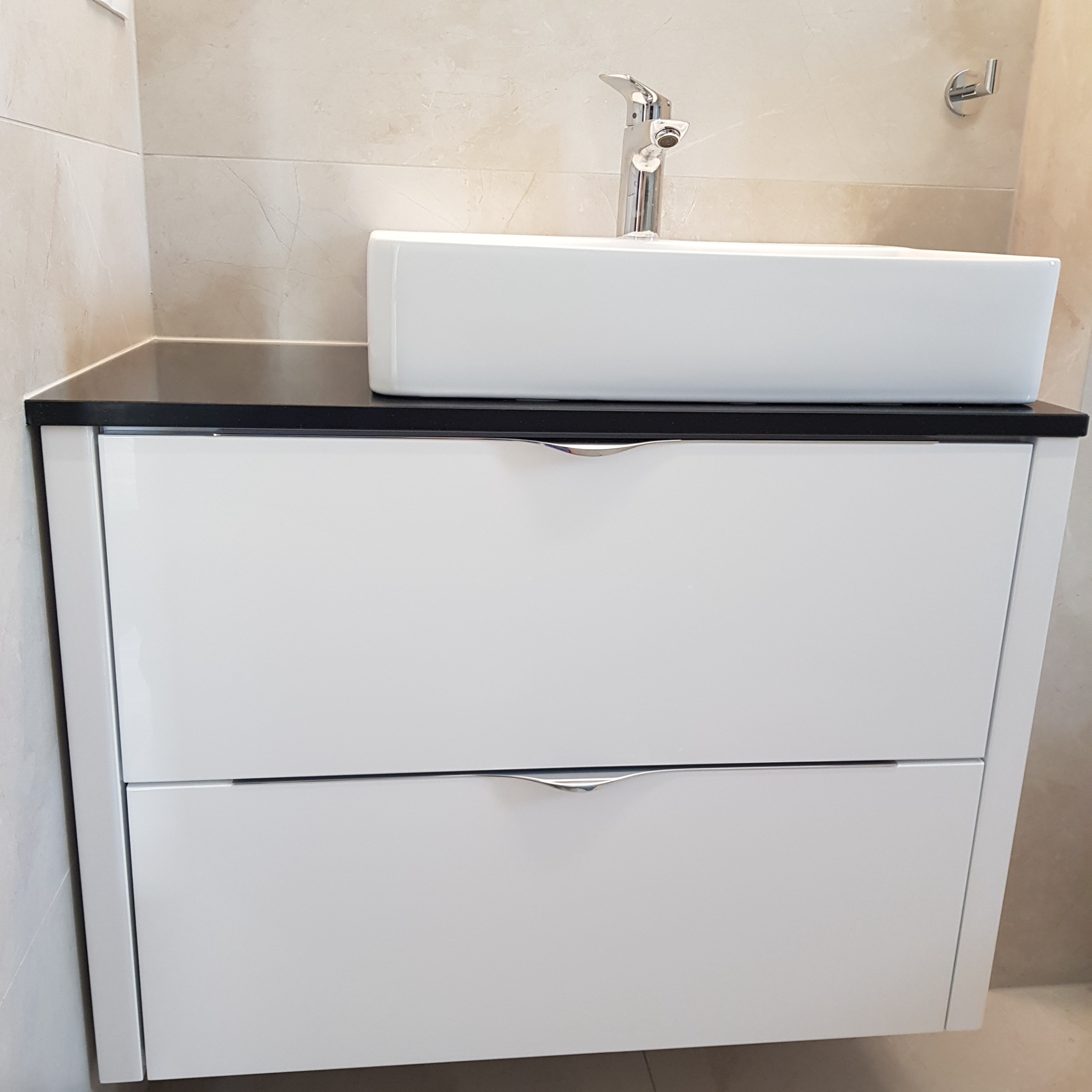 Biała szafka łazienkowa z dwiema szufladami i czarnym blatem, na którym stoi prostokątna umywalka i chromowana bateria, na tle beżowych płytek.