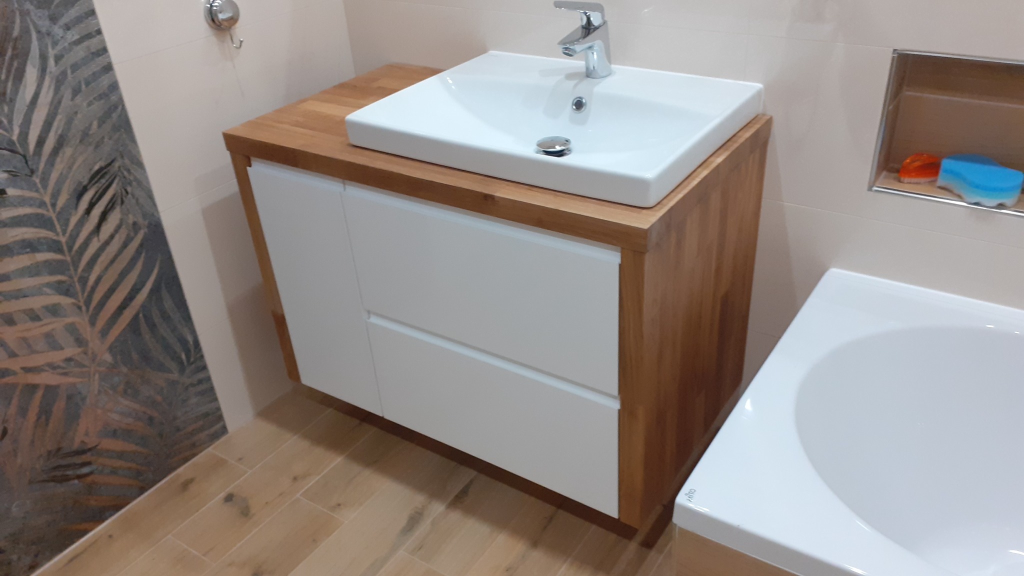 Łazienka z drewnianym blatem pod umywalką i białymi szufladami, płytki podłogowe imitujące drewno, dekoracyjne płytki ścienne z motywem liści.