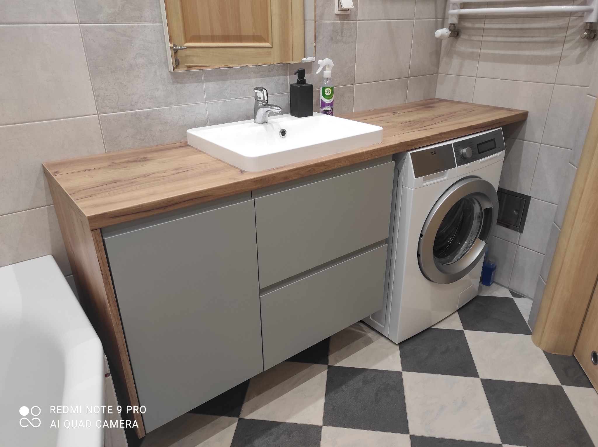 Łazienka z umywalką nablatową i szarą szafką z dwiema szufladami pod blatem z drewnopodobnego laminatu, obok pralka, podłoga w czarno-białą szachownicę.