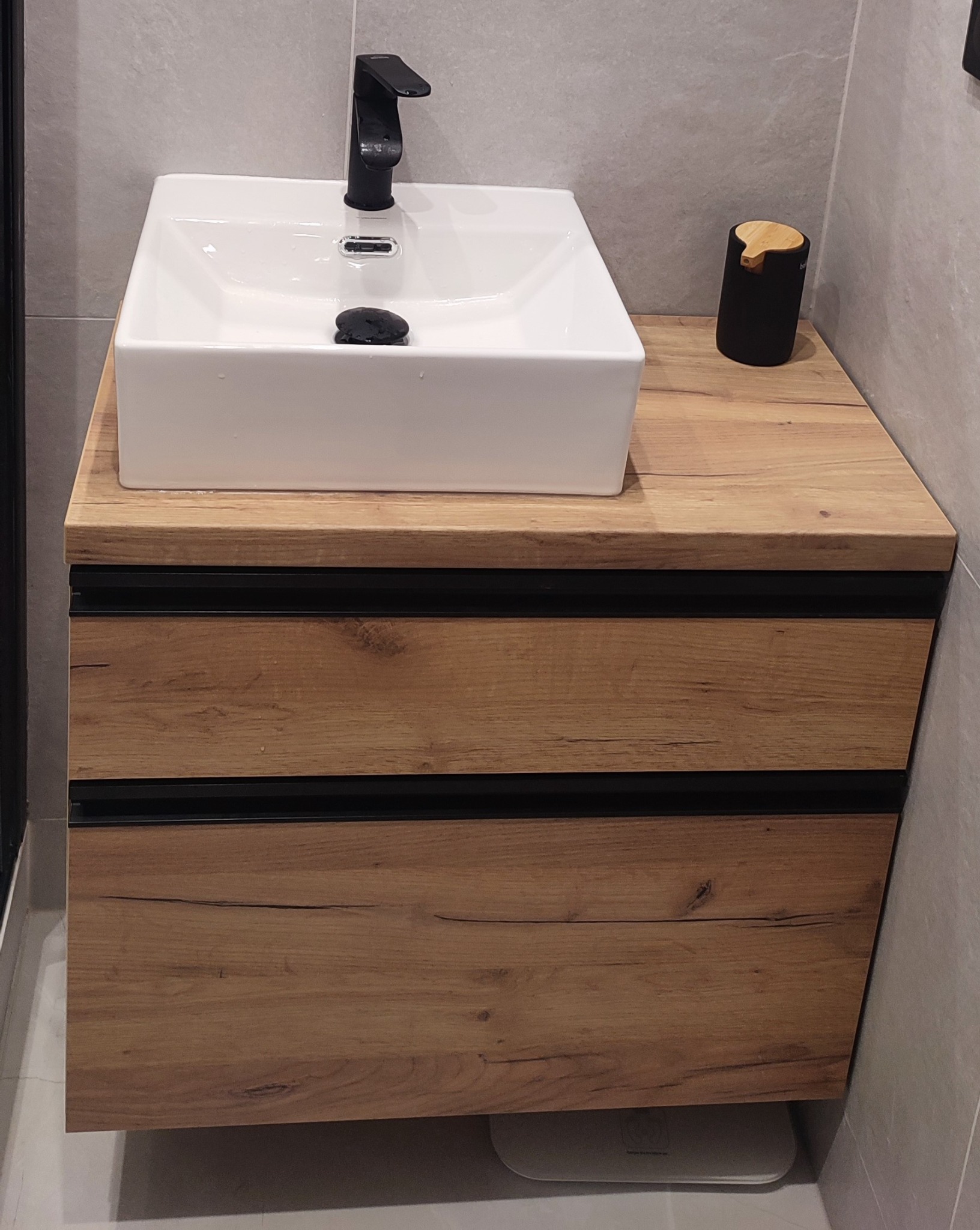 Nowoczesna szafka łazienkowa z drewnianymi szufladami i białą, kwadratową umywalką nablatową oraz czarną armaturą, uzupełniona czarnym dozownikiem na mydło z bambusowym akcentem.