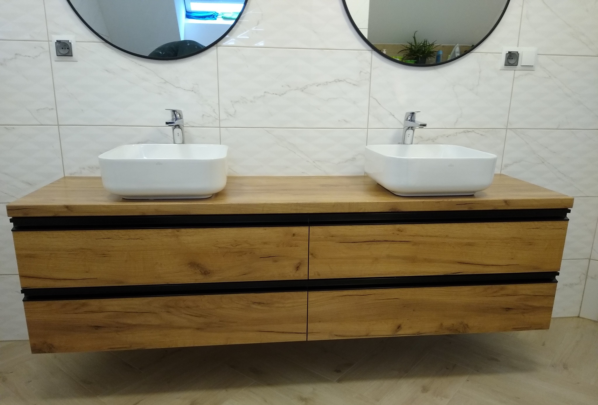 Nowoczesna łazienka z podwójną umywalką nablatową na drewnianej szafce z szufladami i czarnymi akcentami, owalne lustra wiszące na marmurowej ścianie.