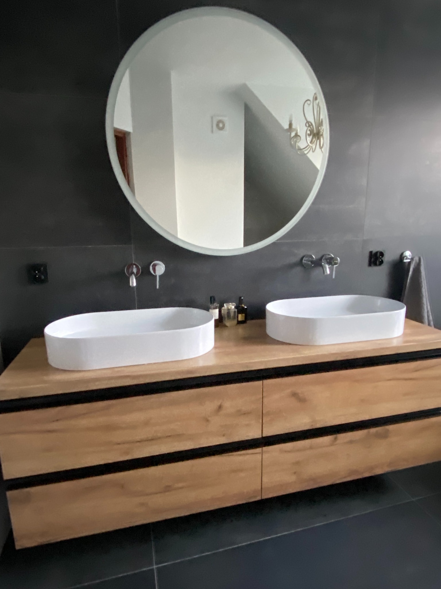 Nowoczesna łazienka z podwójną umywalką nablatową, drewniana szafka z czarnymi akcentami, okrągłe lustro i szara ściana.
