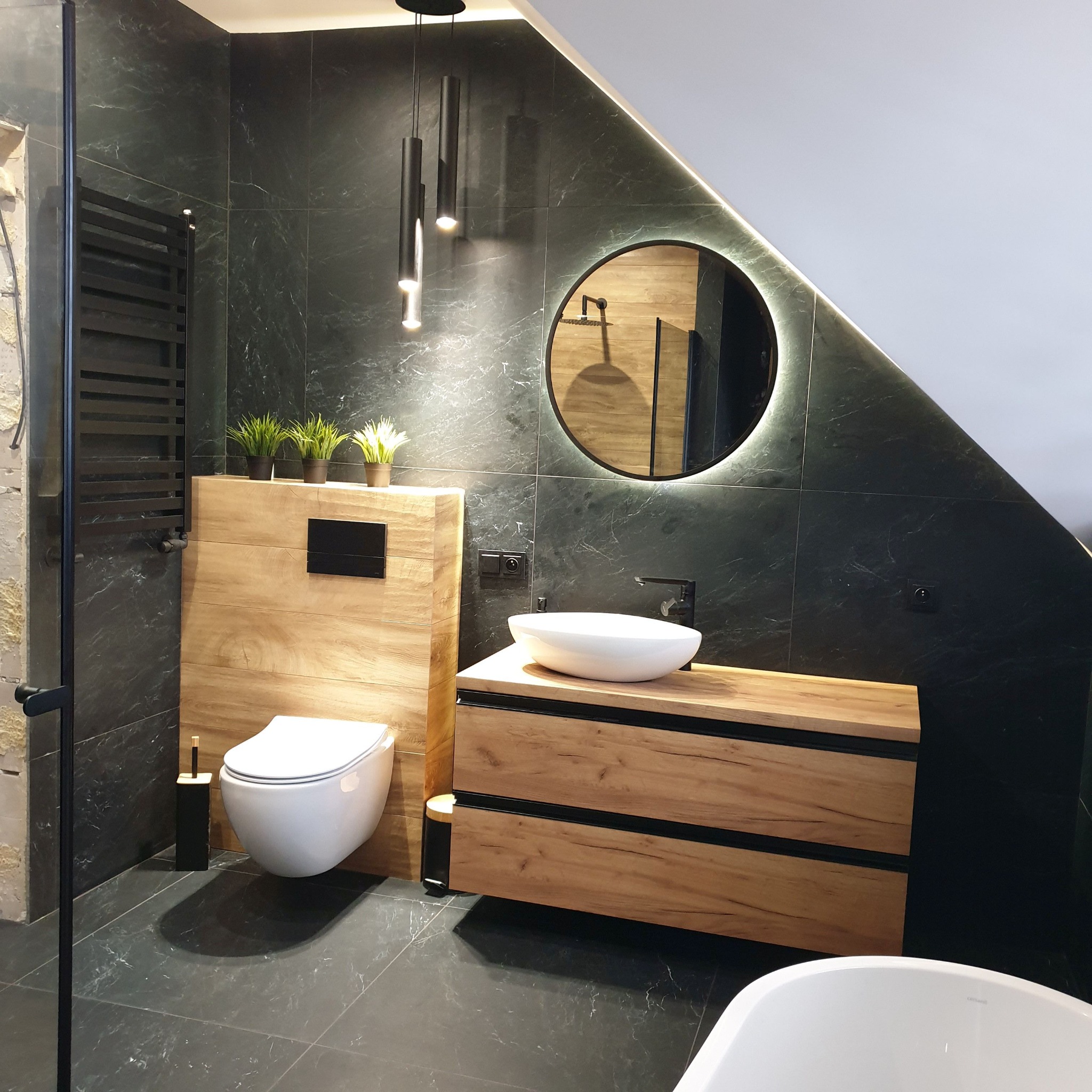 Nowoczesna łazienka z drewnianą szafką pod umywalkę, okrągłym lustrem z podświetleniem LED i czarnymi płytkami imitującymi marmur.