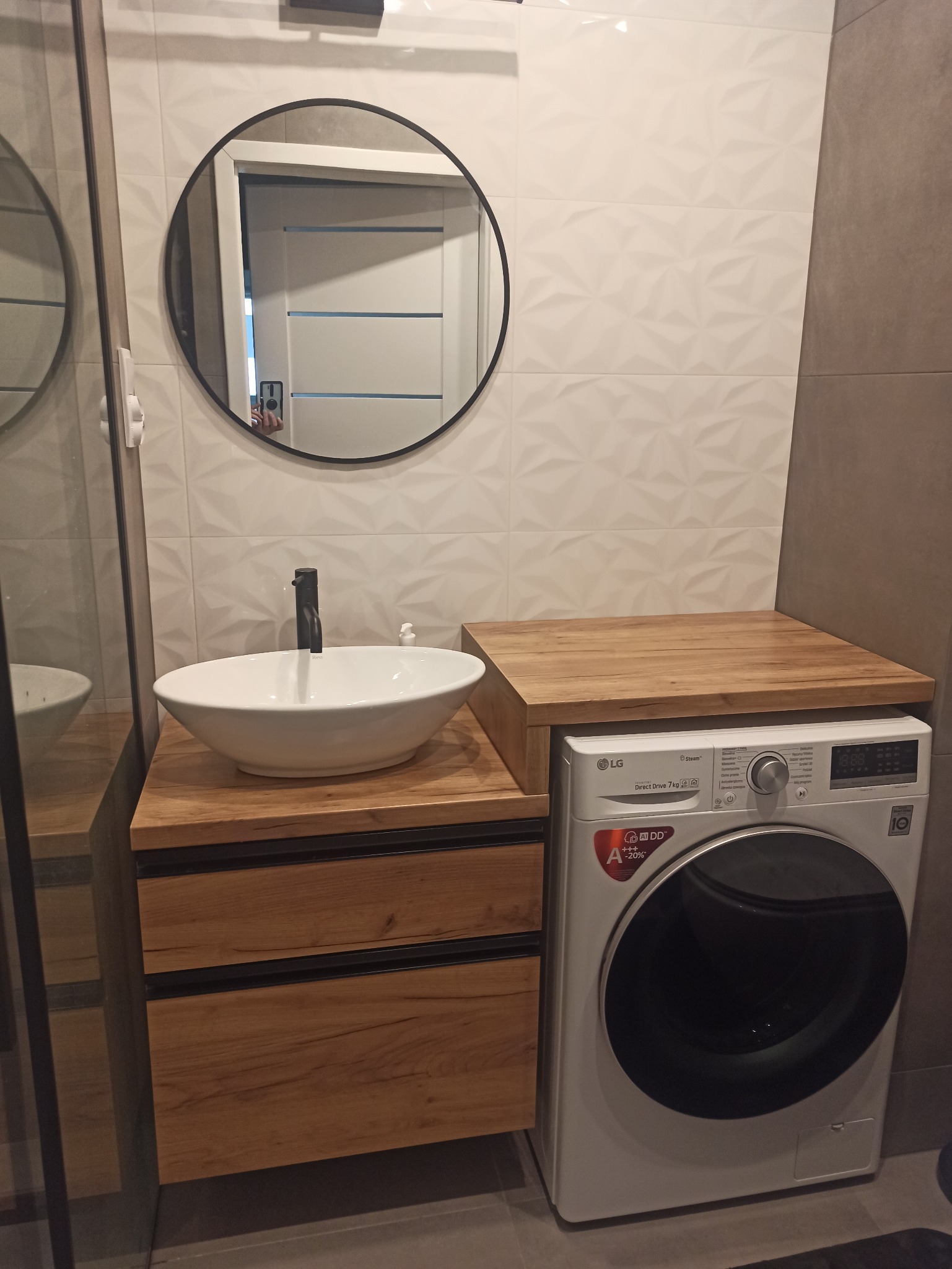 Nowoczesna łazienka z drewnianą szafką pod umywalkę nablatową i blatem nad pralką LG, okrągłe lustro z czarną ramą, białe płytki 3D na ścianie.