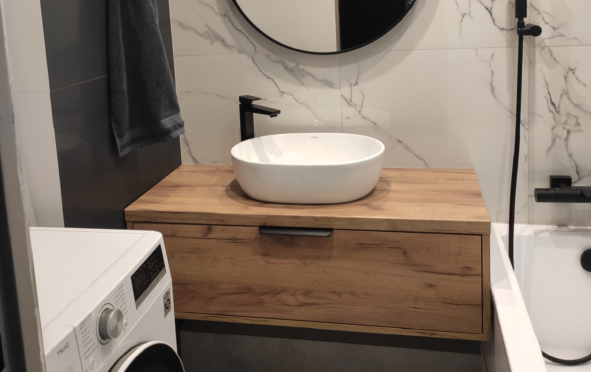 Biała umywalka nablatowa Calani na drewnianej szafce łazienkowej z szufladą, czarna bateria, marmurowe płytki na ścianie, pralka LG z boku.