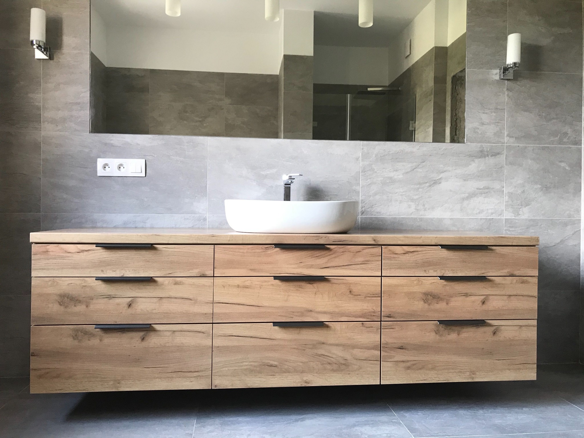 Łazienka z drewnianą szafką pod umywalkę nablatową, dziewięć szuflad z czarnymi uchwytami, szare płytki na ścianie i podłodze, duże lustro, oświetlenie punktowe.