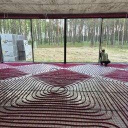 MITRA - Instalacja ogrzewania podłogowego z rurkami w kształcie spirali na folii, widok na las przez duże okno, bloczki Austotherm w rogu. Praca w trakcie realizacji.