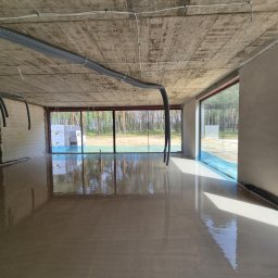 MITRA - Świeżo wylana, mokra posadzka w surowym wnętrzu z dużymi oknami, przez które widać las. Na suficie widoczne szare rury wentylacyjne.