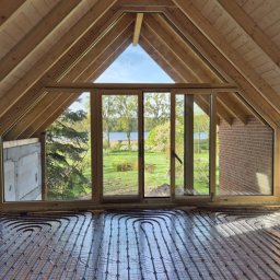 MITRA - Montaż ogrzewania podłogowego w nowym domu z widokiem na jezioro. Rury grzewcze ułożone na folii, drewniana konstrukcja dachu i duże okno przesuwne.