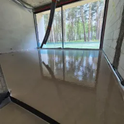 Świeżo wylana, gładka posadzka w surowym wnętrzu z widokiem na las przez duże okno. W tle widoczne rury instalacyjne pod sufitem.