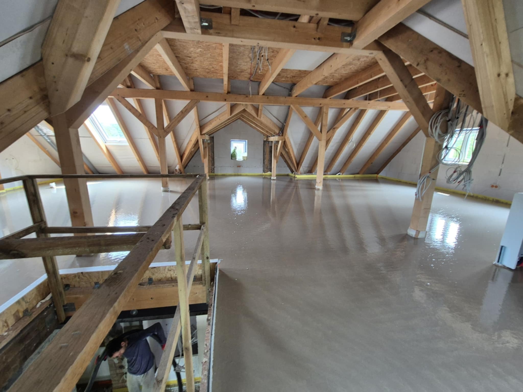Świeżo wylana, mokra posadzka na poddaszu z drewnianą konstrukcją więźby dachowej. Widoczne okna dachowe i pracownik w tle. Żółta taśma na obrzeżach.