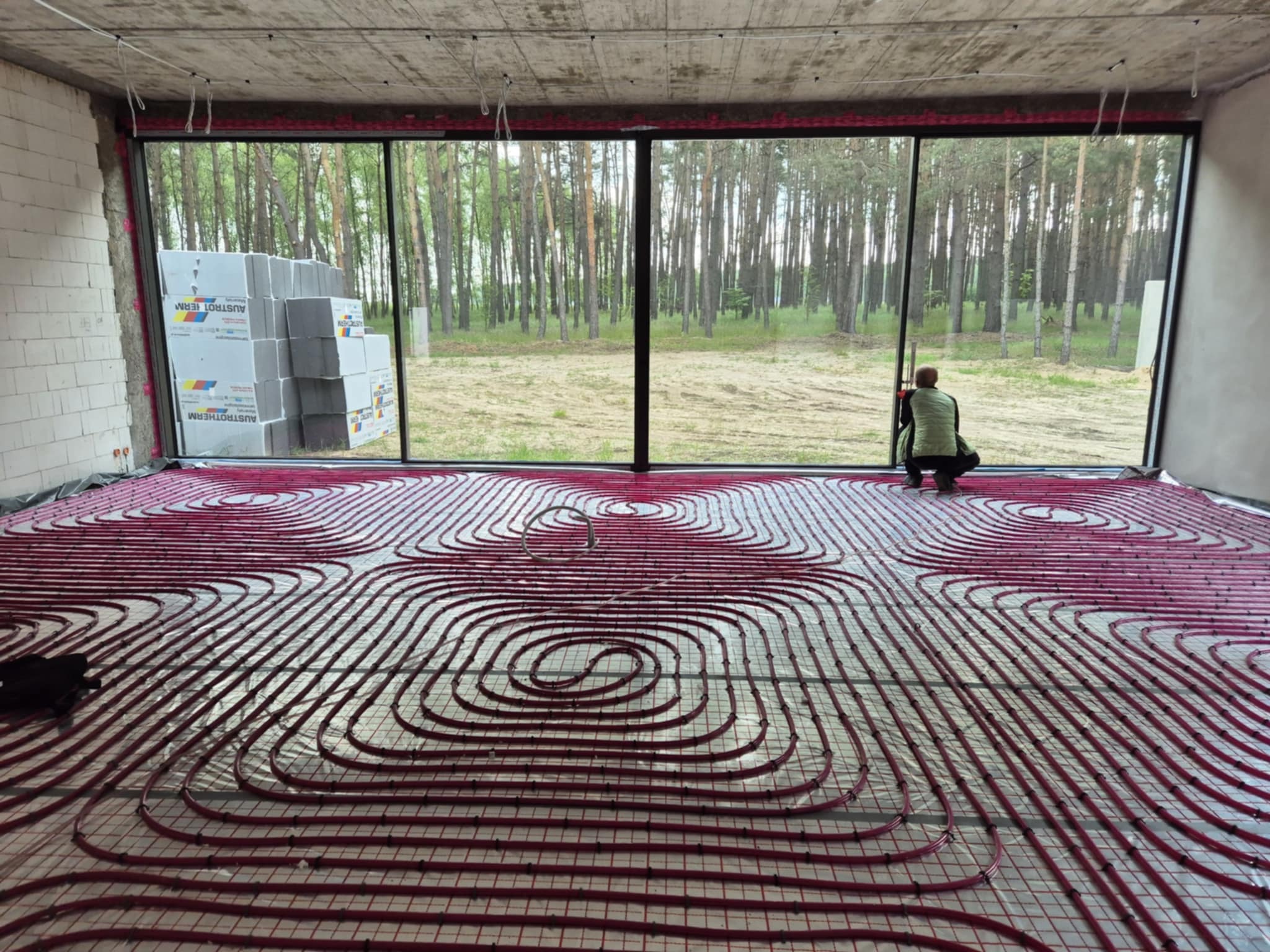 Instalacja ogrzewania podłogowego z rurkami w kształcie spirali na folii, widok na las przez duże okno, bloczki Austotherm w rogu. Praca w trakcie realizacji.