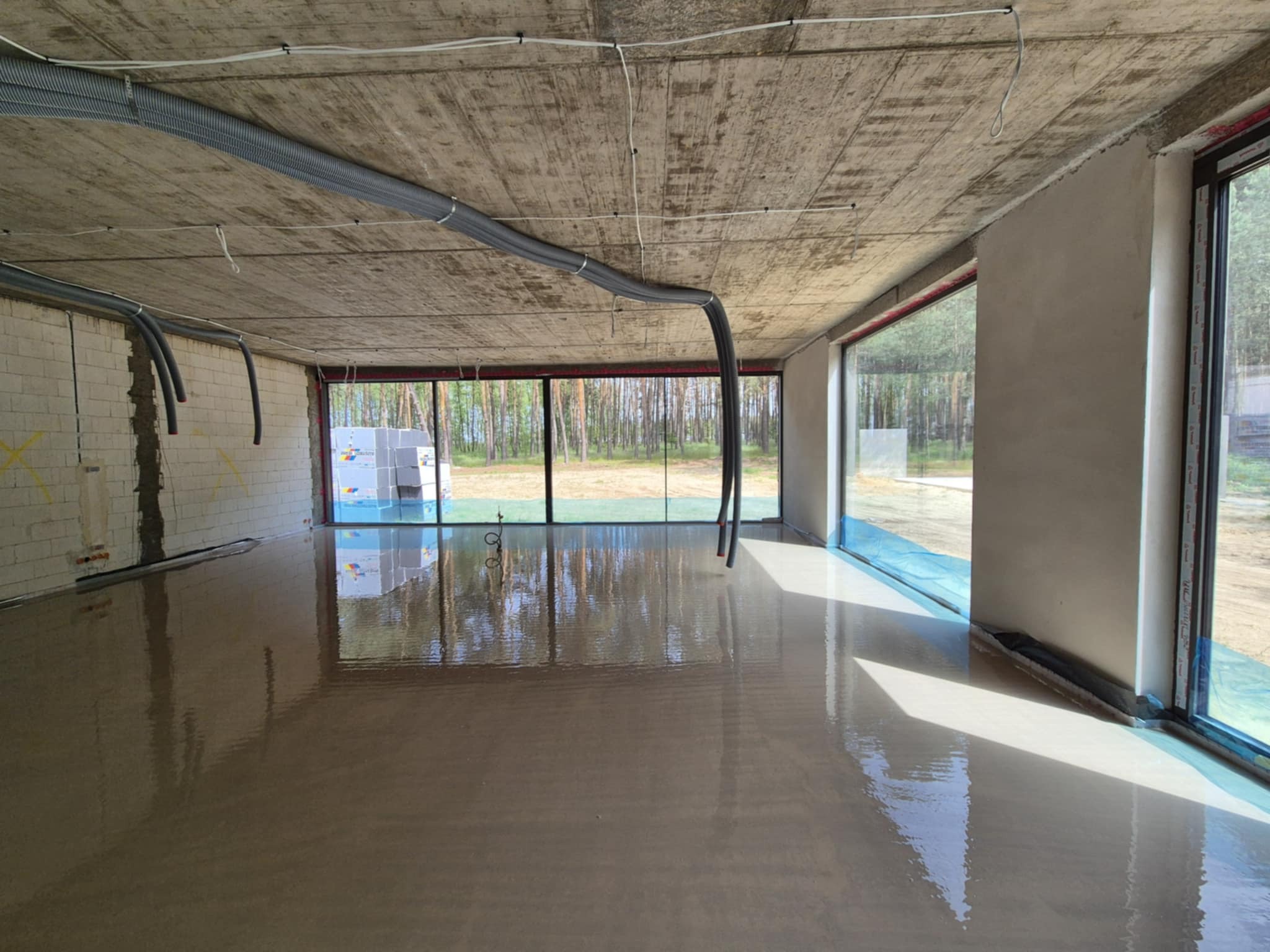 Świeżo wylana, mokra posadzka w surowym wnętrzu z dużymi oknami, przez które widać las. Na suficie widoczne szare rury wentylacyjne.