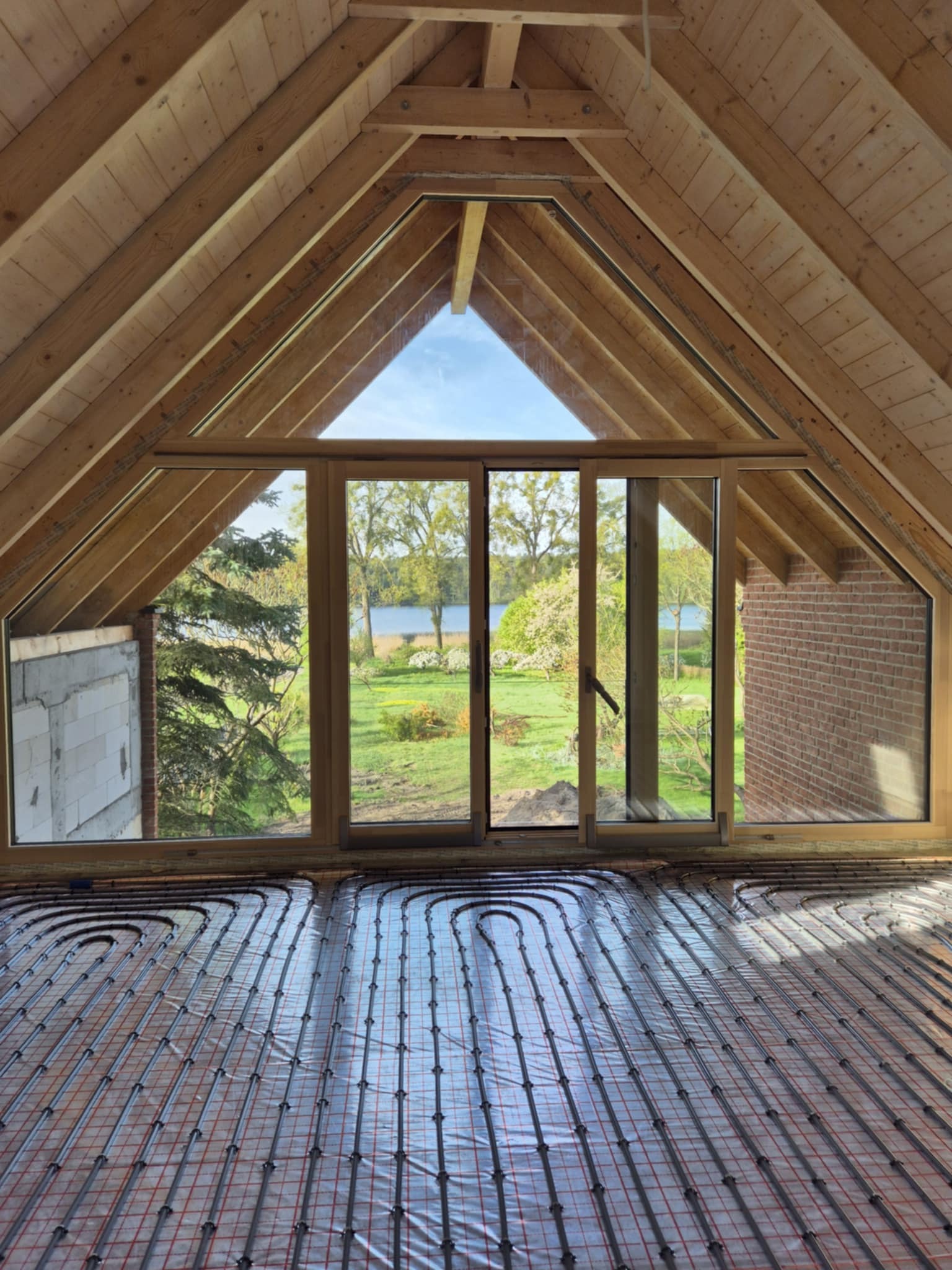Montaż ogrzewania podłogowego w nowym domu z widokiem na jezioro. Rury grzewcze ułożone na folii, drewniana konstrukcja dachu i duże okno przesuwne.