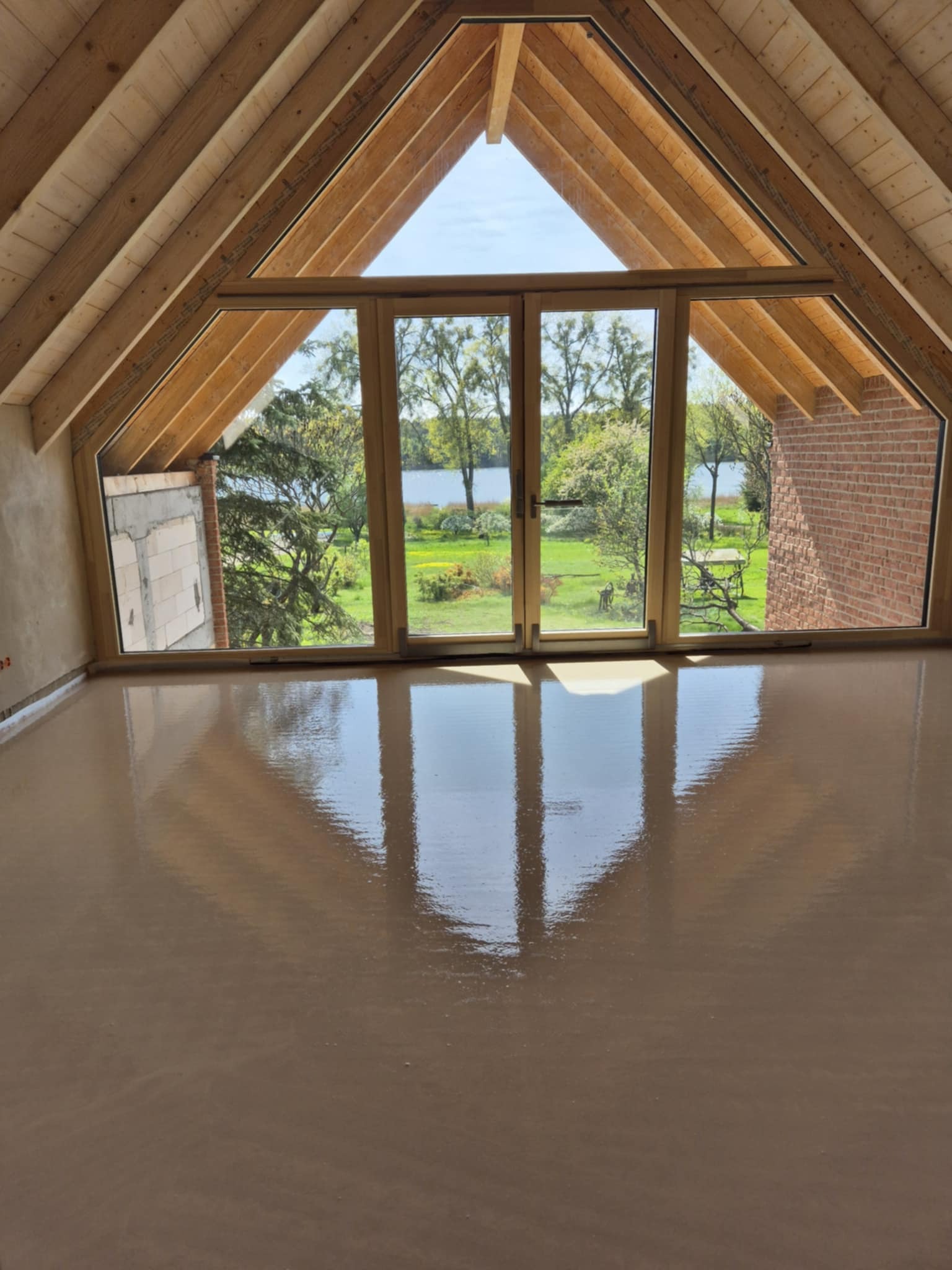 Świeżo wylana, gładka, beżowa posadzka w jasnym pokoju na poddaszu z drewnianą konstrukcją dachu i dużym oknem z widokiem na zieleń i wodę.