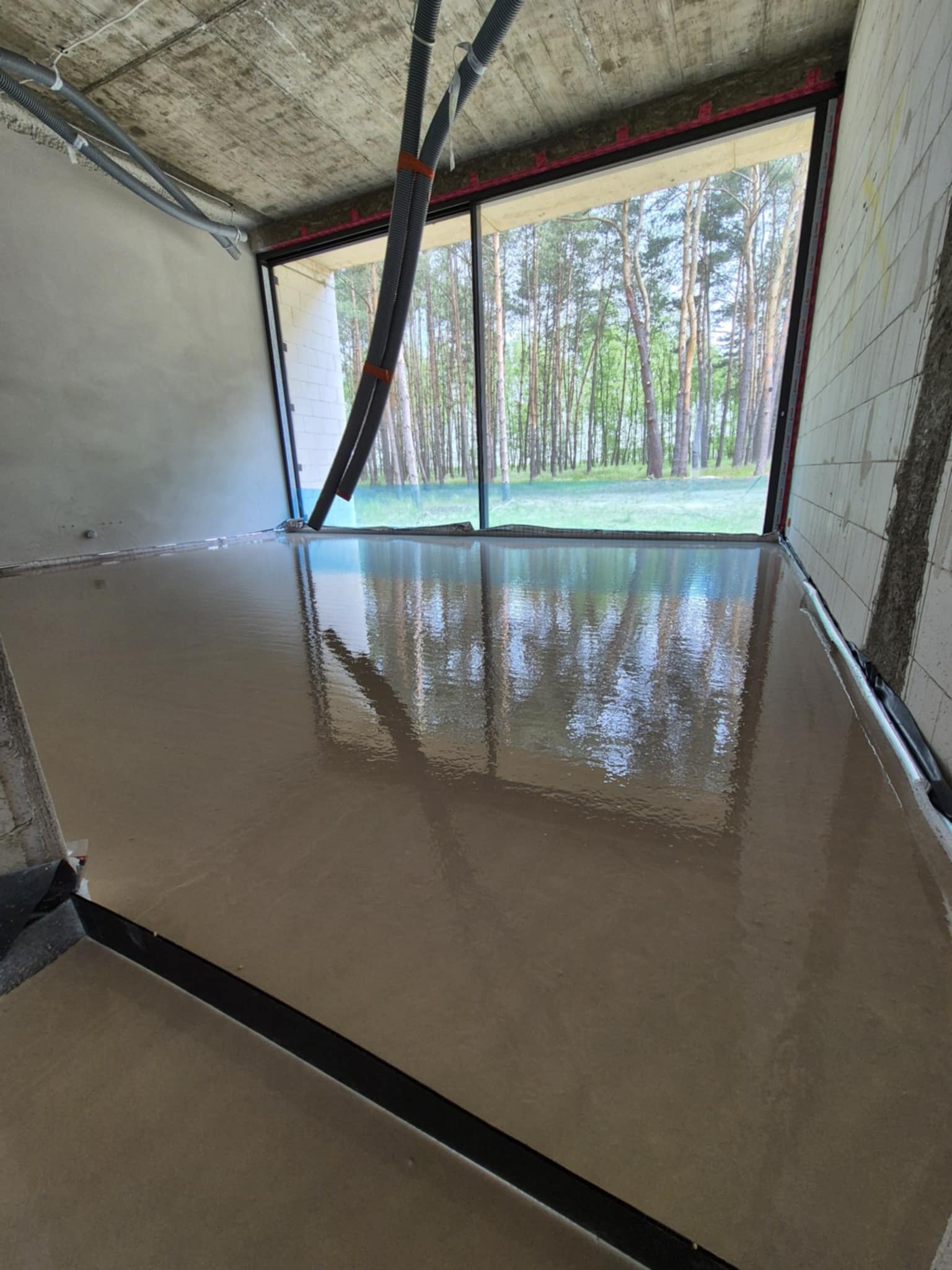 Świeżo wylana, gładka posadzka w surowym wnętrzu z widokiem na las przez duże okno. W tle widoczne rury instalacyjne pod sufitem.