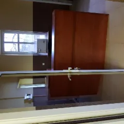 Wnętrze pomieszczenia z dwukolorową ścianą (beż i brąz), oknem z widokiem na drzewa i brązową szafką, widziane przez szklaną ścianę lub drzwi z białą ramą.