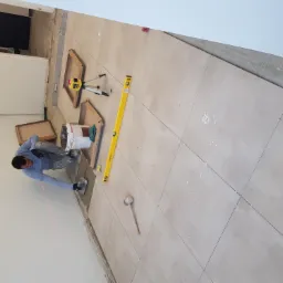 Układanie beżowych płytek podłogowych w pomieszczeniu, widoczny pracownik z kielnią, poziomica laserowa i tradycyjna, oraz krzyżyki dystansowe między płytkami.