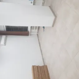 Puste pomieszczenie z jasnymi, kwadratowymi płytkami podłogowymi, klimatyzatorem na ścianie, brązową roletą w oknie i stosem paneli podłogowych w rogu. W tle widoczna wiertarka przy ścianie.