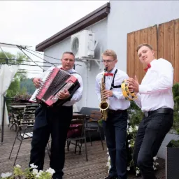 Trzech muzyków w białych koszulach i muchach gra na akordeonie, saksofonie i tamburynie na drewnianym tarasie z widokiem na zieleń.