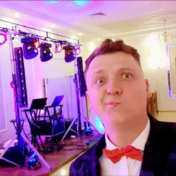 Mężczyzna w muszce robi selfie na tle rozstawionego sprzętu nagłośnieniowego i oświetleniowego w sali bankietowej z dekoracjami.