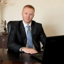 Młody mężczyzna w garniturze siedzi za biurkiem z laptopem, w tle wazon z suchymi kwiatami i statuetka Temidy.