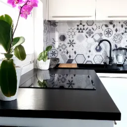 Nowoczesna kuchnia z czarnym blatem drewnianym, białymi szafkami i geometrycznym wzorem na ścianie, z elementami dekoracyjnymi jak storczyk i pianino.