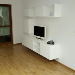 Nowoczesny salon z białymi, wiszącymi szafkami i półkami pod telewizorem, drewniana podłoga, minimalistyczny wystrój, trzy szare świece na szafce.