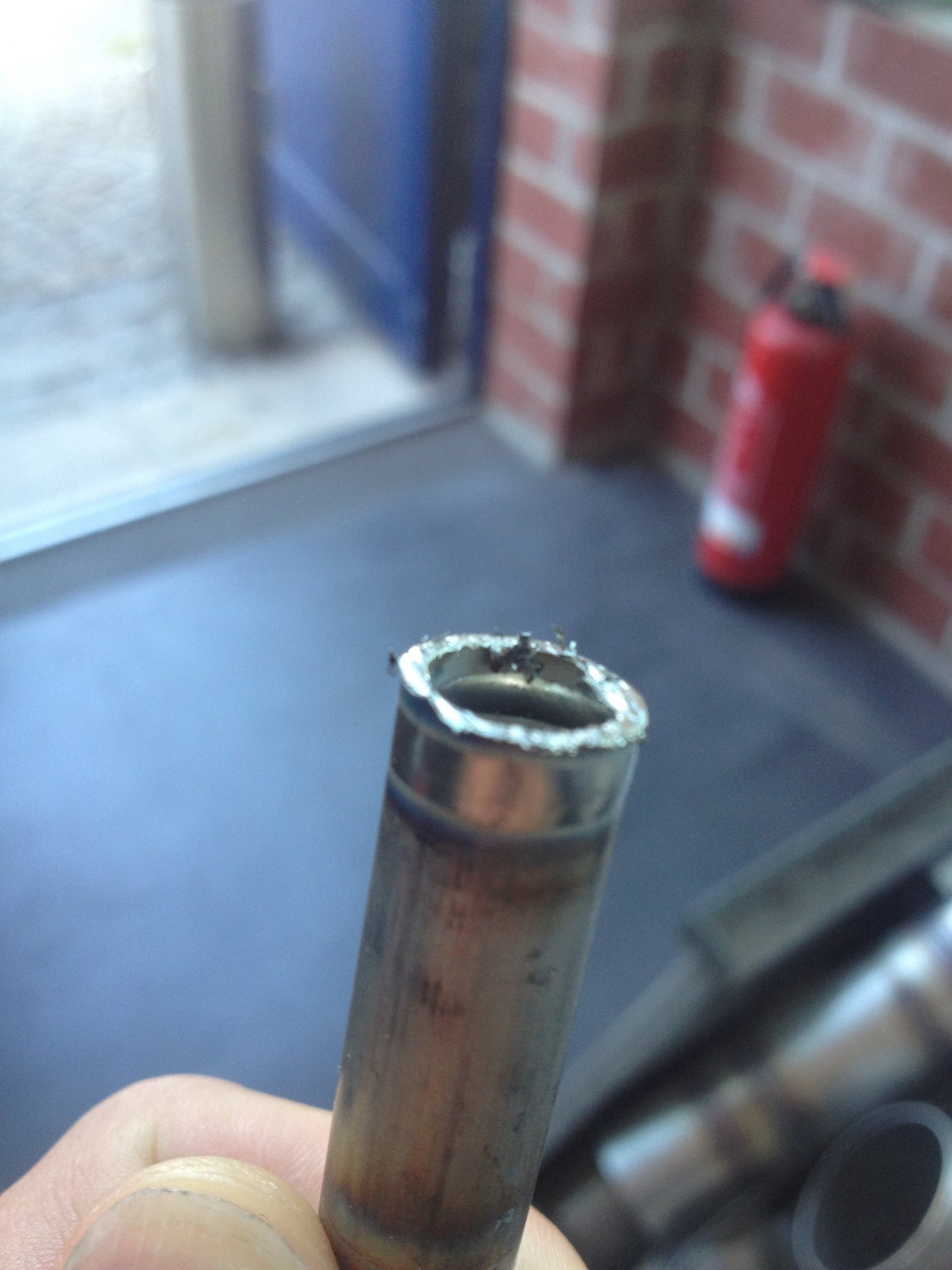 Metalowa rurka z nierównym, poszarpanym brzegiem po obróbce, trzymana w dłoni, widoczne ślady przypaleń i zadziory, w tle ceglana ściana i gaśnica.