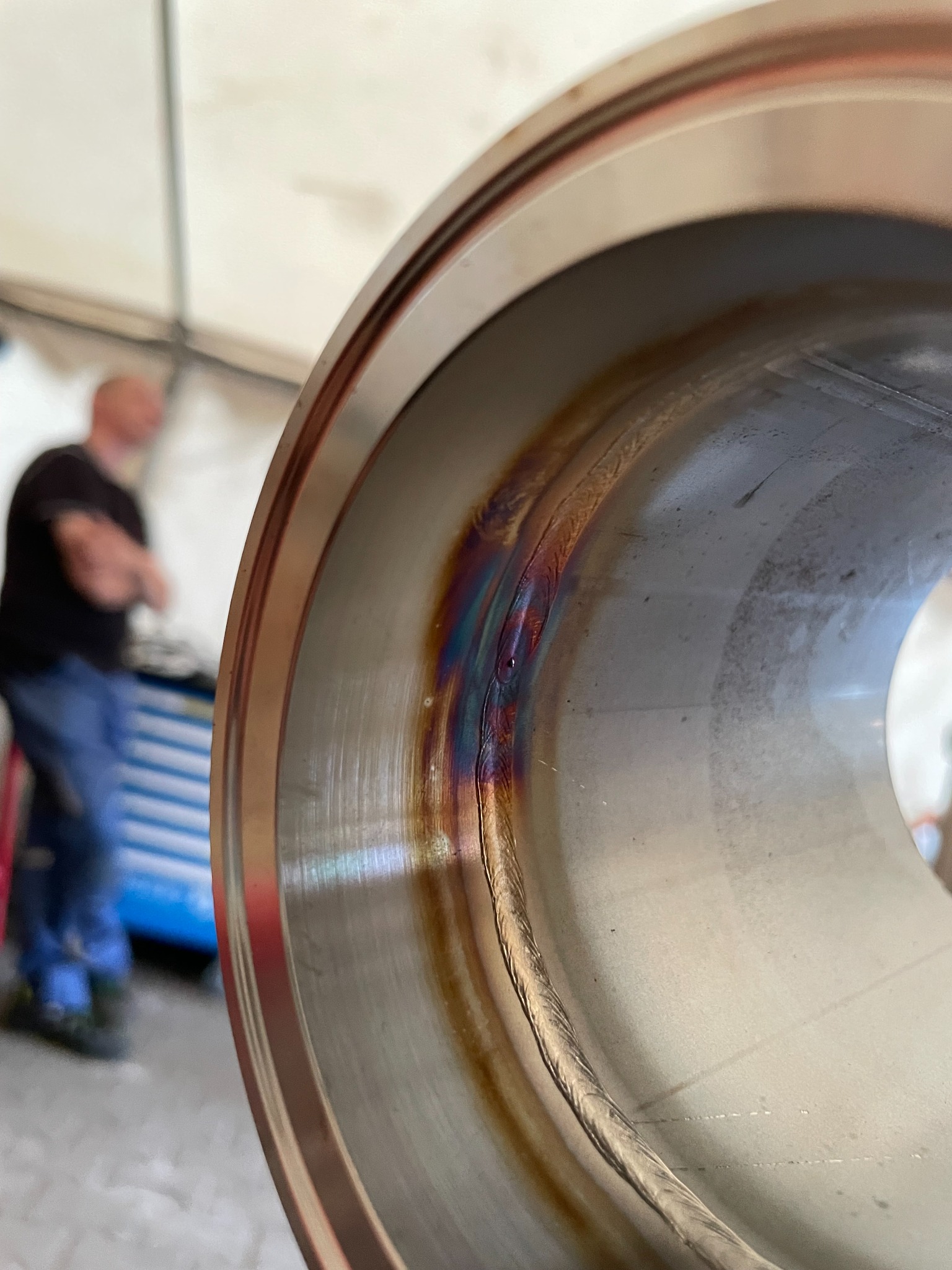 Wnętrze metalowej rury z widocznym spoiwem po procesie łączenia metali, tęczowe zabarwienie w miejscu spawu, w tle rozmyta sylwetka osoby i warsztat.