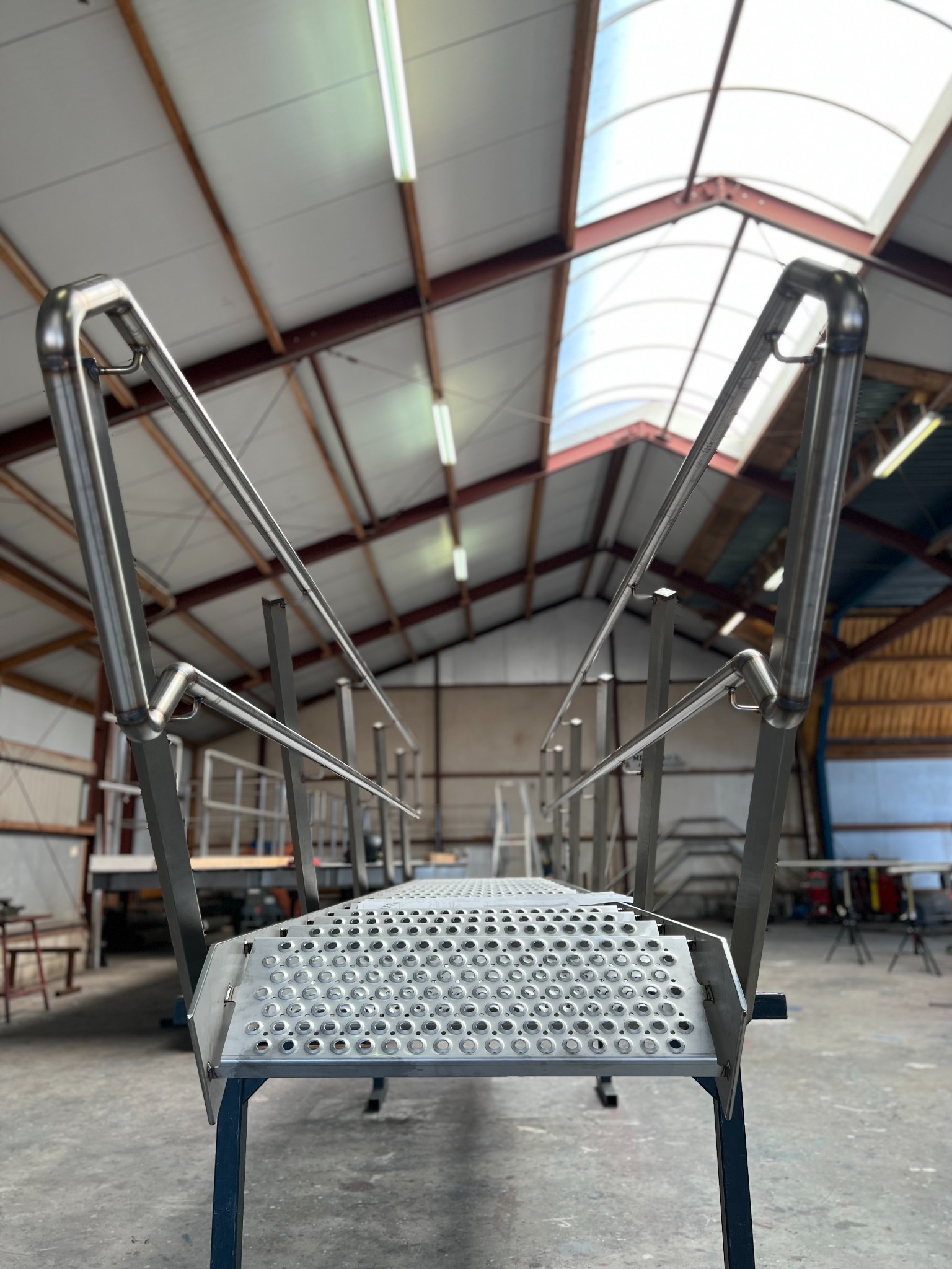 Metalowe schody z perforowanymi stopniami i poręczami, wykonane ze stali nierdzewnej, w hali produkcyjnej, widok z dołu, oświetlenie jarzeniowe.