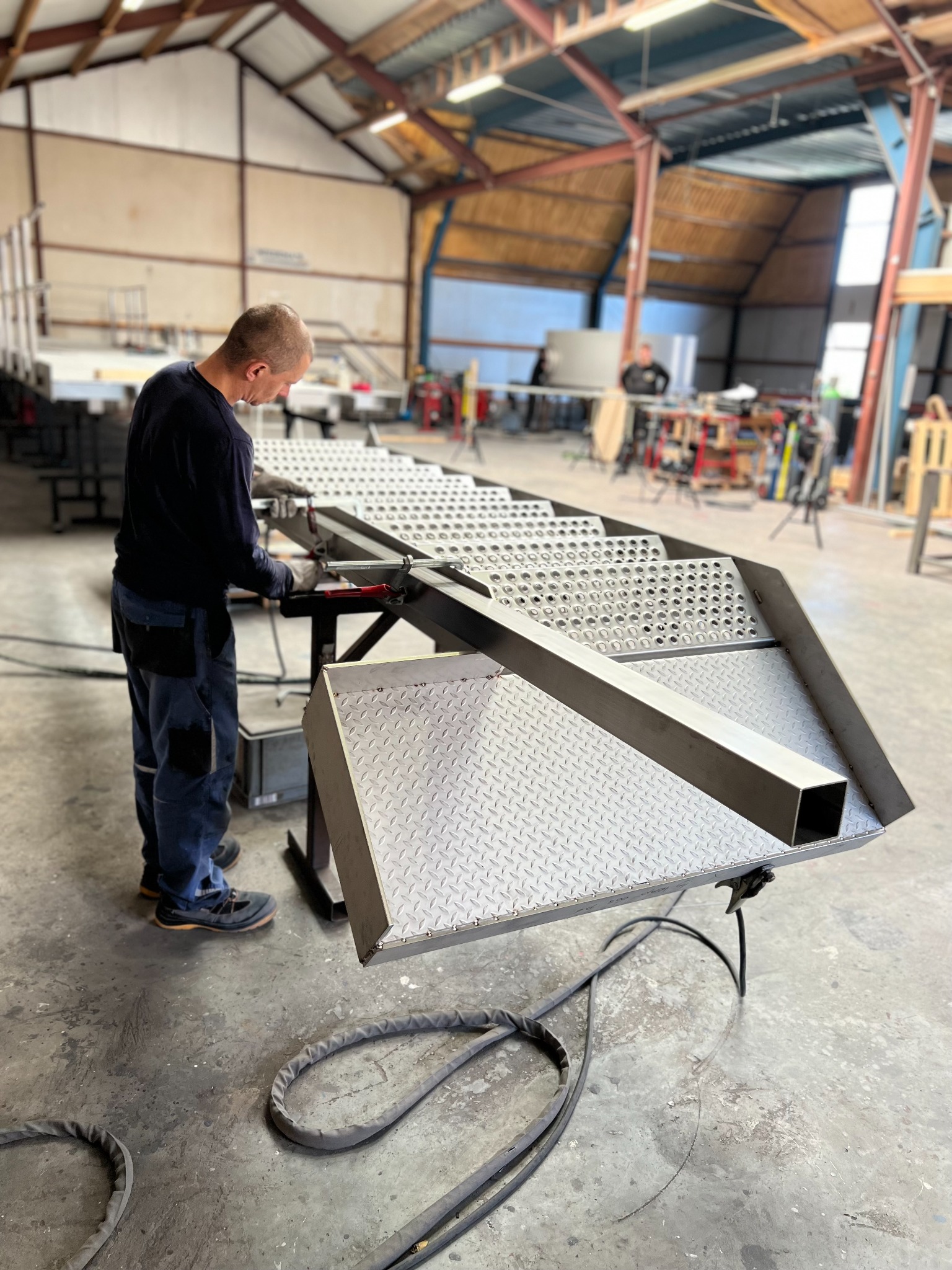 Pracownik w warsztacie łączy metalowe elementy schodów przemysłowych przy użyciu narzędzi spawalniczych; w tle widoczny drugi pracownik i konstrukcja hali.