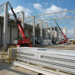 Montaż konstrukcji stalowej hali z użyciem podnośników Manitou i JCB, widoczne płyty warstwowe Kingspan QuadCore, betonowe słupy i stalowe kratownice pod dachem.