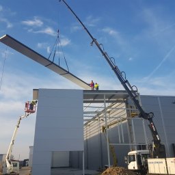 FUH DACHTEX TOMASZ KOWALSKI - Montaż płyty warstwowej na budowie hali. Dwóch pracowników na dachu, jeden w podnośniku koszowym. Dźwig podnosi płytę. Błękitne niebo w tle.