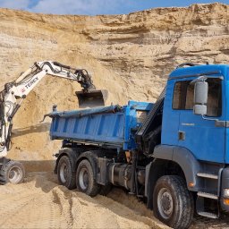 FUH DACHTEX TOMASZ KOWALSKI - Błękitna ciężarówka MAN i biała koparka Terex ładują piasek na tle piaszczystego wzgórza. Widoczny operator koparki i tablica rejestracyjna ciężarówki.