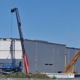 FUH DACHTEX TOMASZ KOWALSKI - Montaż płyty warstwowej na hali przemysłowej z użyciem dźwigu i podnośnika koszowego. Pracownicy w kaskach. Widok na budynek w trakcie prac.