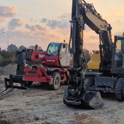 FUH DACHTEX TOMASZ KOWALSKI - Żółta koparka kołowa, czerwony podnośnik teleskopowy Manitou i niebieska wywrotka MAN na placu budowy o zmierzchu. Widoczne ślady opon na piasku.