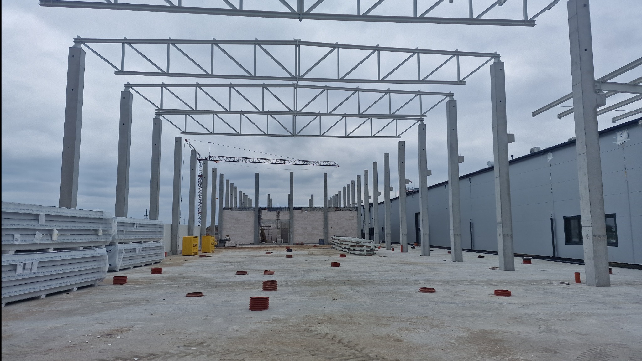 Wnętrze budowanej hali z betonowymi słupami i stalową konstrukcją dachu. W tle widoczny żuraw budowlany i ściany z pustaków. Na podłożu widoczne elementy montażowe.