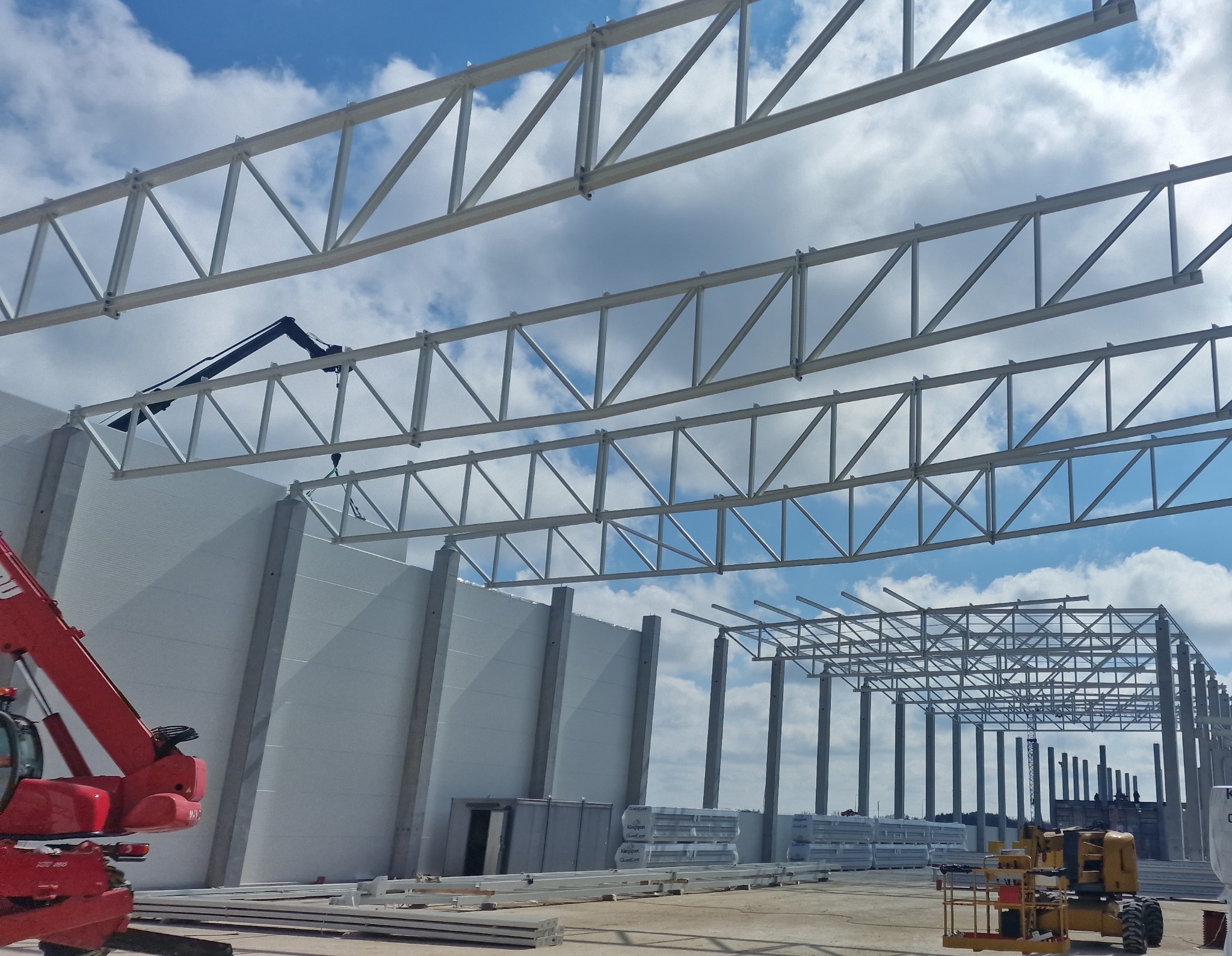 Budowa hali: montaż stalowej konstrukcji nośnej z użyciem dźwigu na tle błękitnego nieba. Widoczne płyty warstwowe i maszyny budowlane.