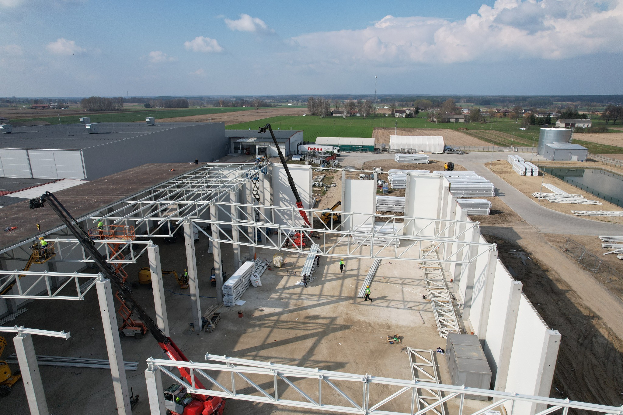 Budowa hali produkcyjnej w toku: montaż stalowej konstrukcji szkieletowej z użyciem dźwigów, widoczne elementy ścian i pracownicy w kaskach, perspektywa z lotu ptaka.