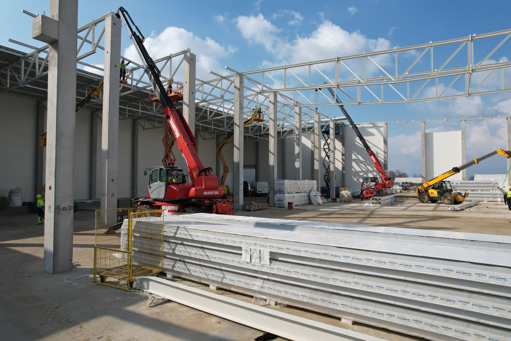Montaż konstrukcji stalowej hali z użyciem podnośników Manitou i JCB, widoczne płyty warstwowe Kingspan QuadCore, betonowe słupy i stalowe kratownice pod dachem.