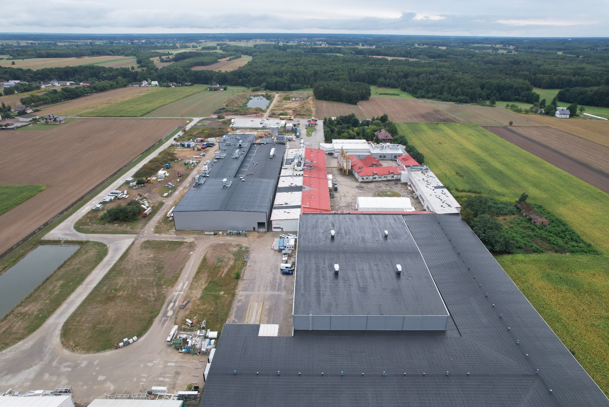 Szeroki kadr z lotu ptaka na kompleks hal przemysłowych z ciemnymi dachami, otoczonych polami uprawnymi i lasami. Widoczne zbiorniki wodne i drogi dojazdowe.