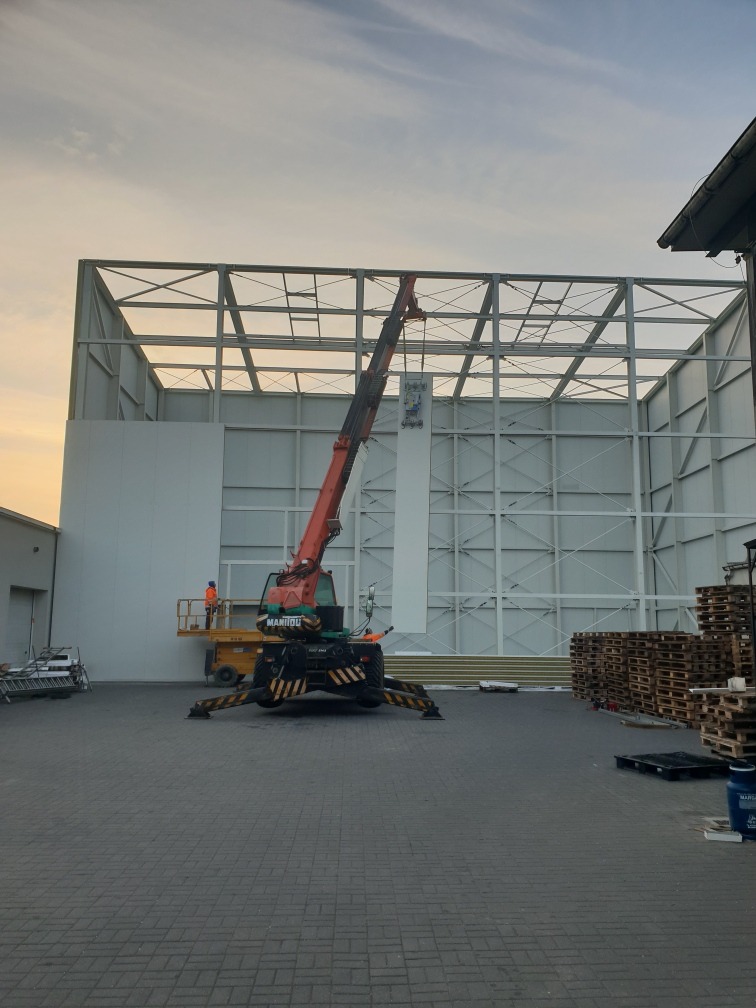Montaż hali z płyt warstwowych: widok na konstrukcję stalową, częściowo obudowaną, z dźwigiem Manitou w trakcie pracy i paletami w tle, przy zachmurzonym niebie.