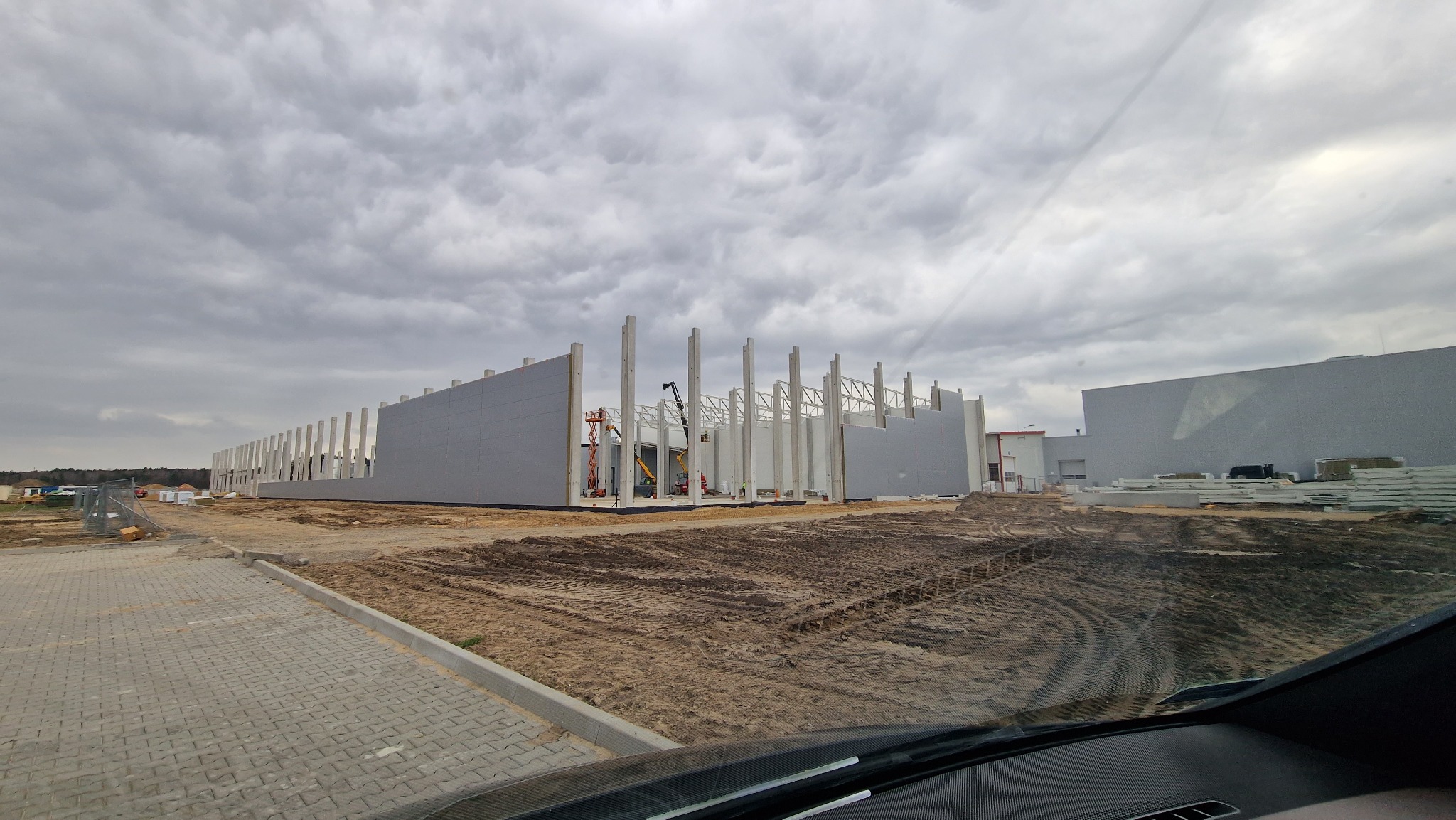 Budowa hali w toku: widoczne betonowe słupy, ściany z płyt warstwowych, żurawie. Pochmurne niebo. Fragment kostki brukowej i wnętrza pojazdu w kadrze.