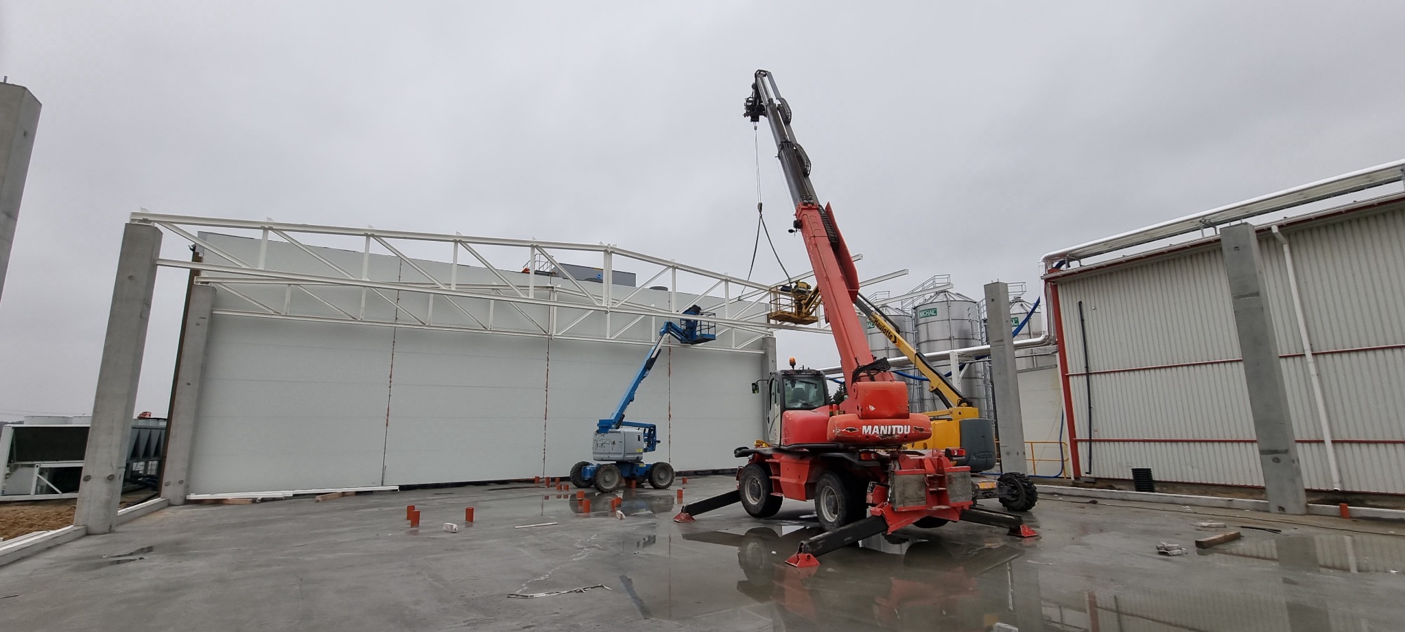 Montaż hali stalowej z użyciem podnośników koszowych. Widoczne elementy konstrukcyjne, ściany z płyt warstwowych i silosy w tle. Prace w toku, pochmurny dzień.